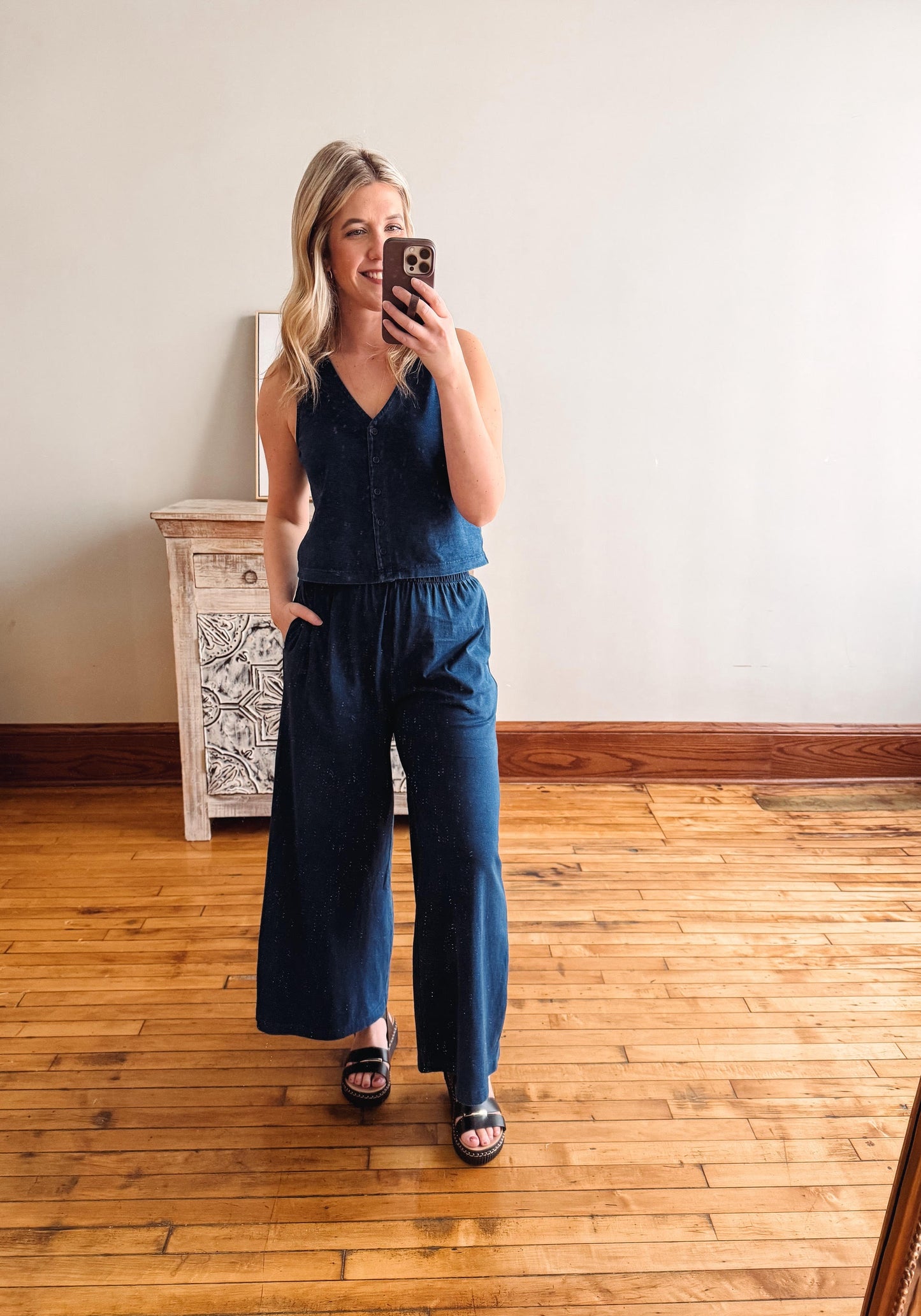 Woman taking a mirror selfie wearing a blue jumpsuit in a room with wooden flooring and a white wall.