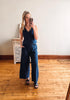 Woman taking a mirror selfie wearing a blue jumpsuit in a room with wooden flooring and a white wall.