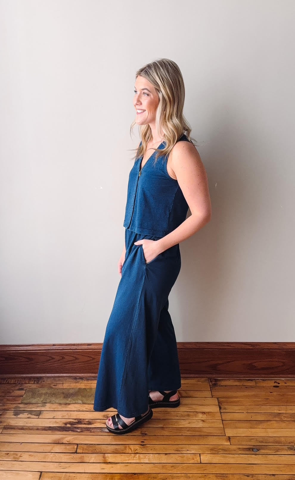 Woman wearing a blue jumpsuit standing against a white wall on a wooden floor.