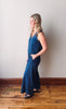 Woman wearing a blue jumpsuit standing against a white wall on a wooden floor.