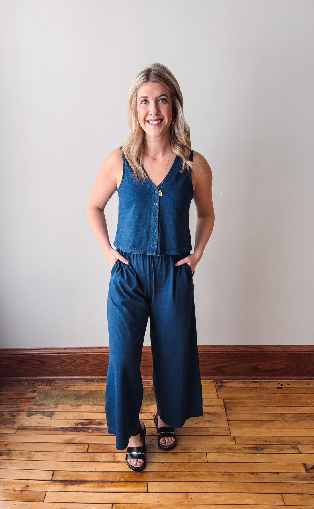 Woman wearing a blue denim jumpsuit standing on a wooden floor with a plain wall background
