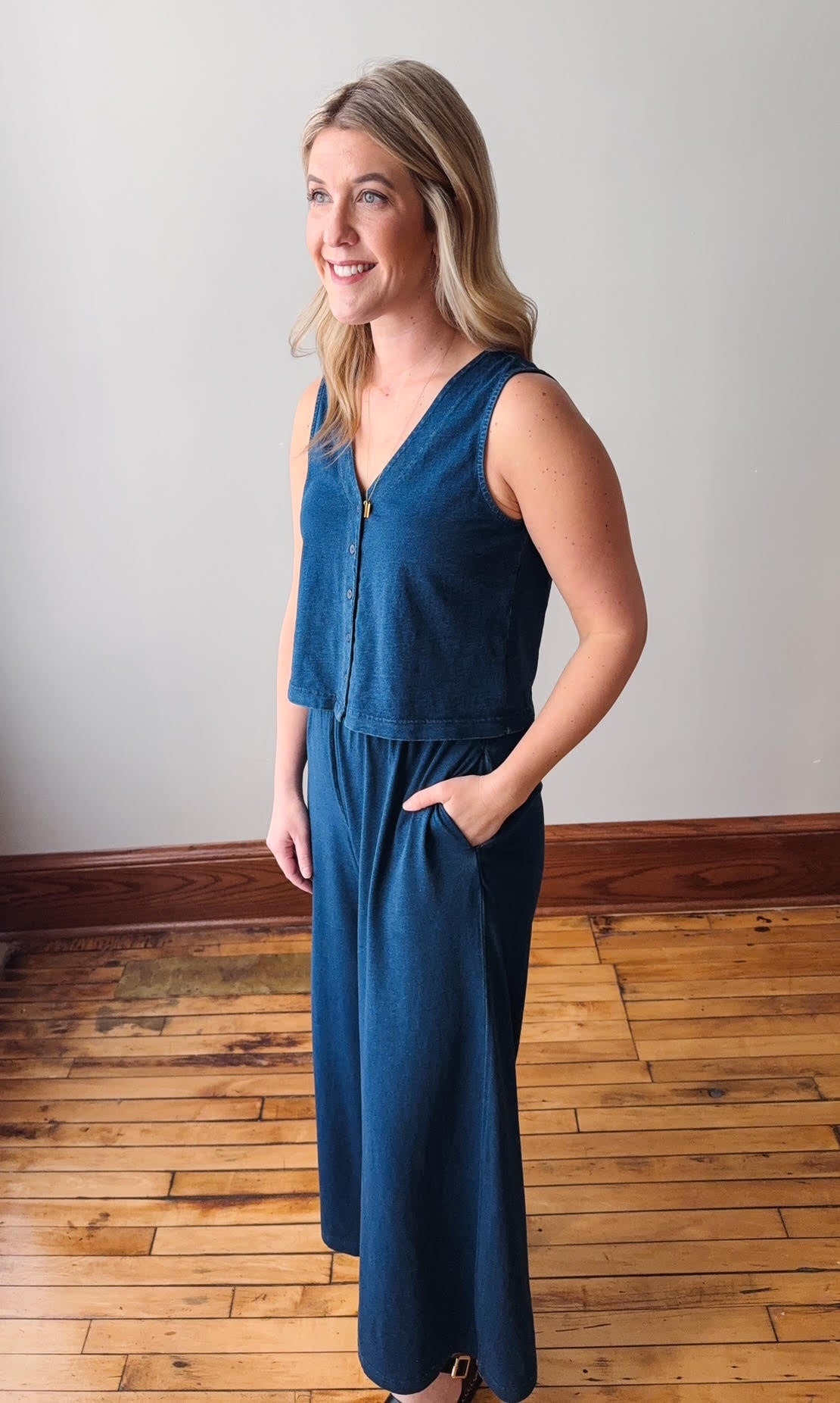 Woman wearing a blue jumpsuit standing on a wooden floor with a white wall background