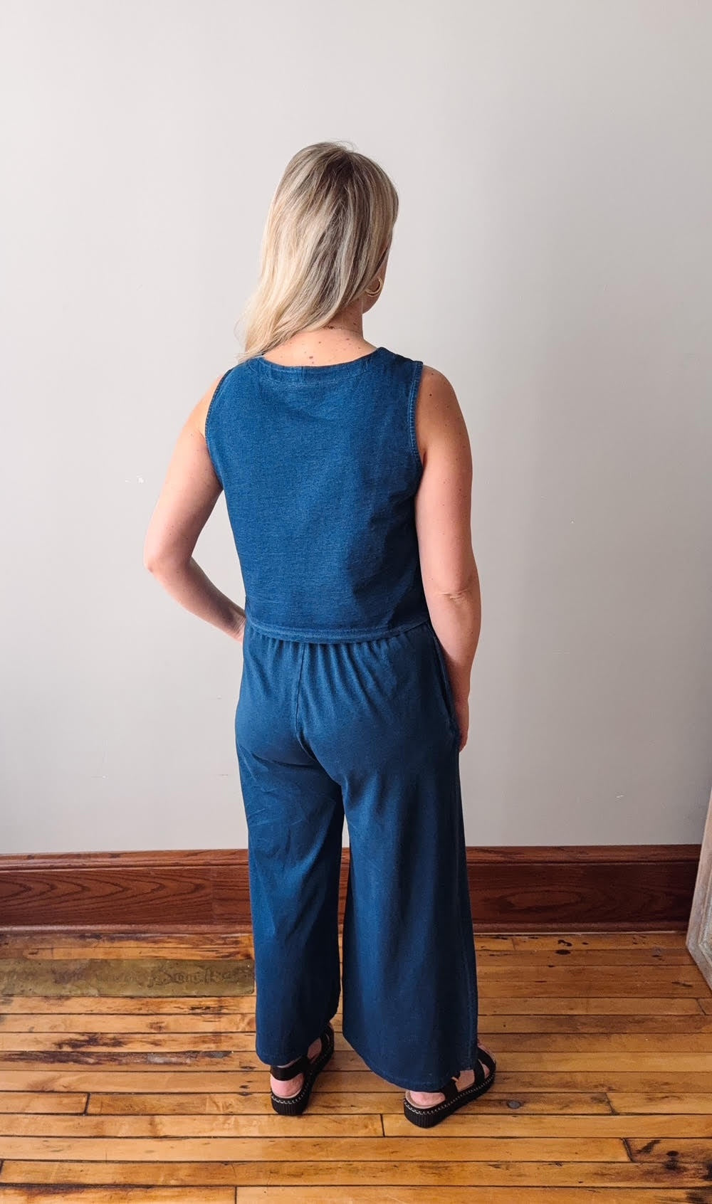 Woman wearing a blue jumpsuit standing on a wooden floor with a plain wall background