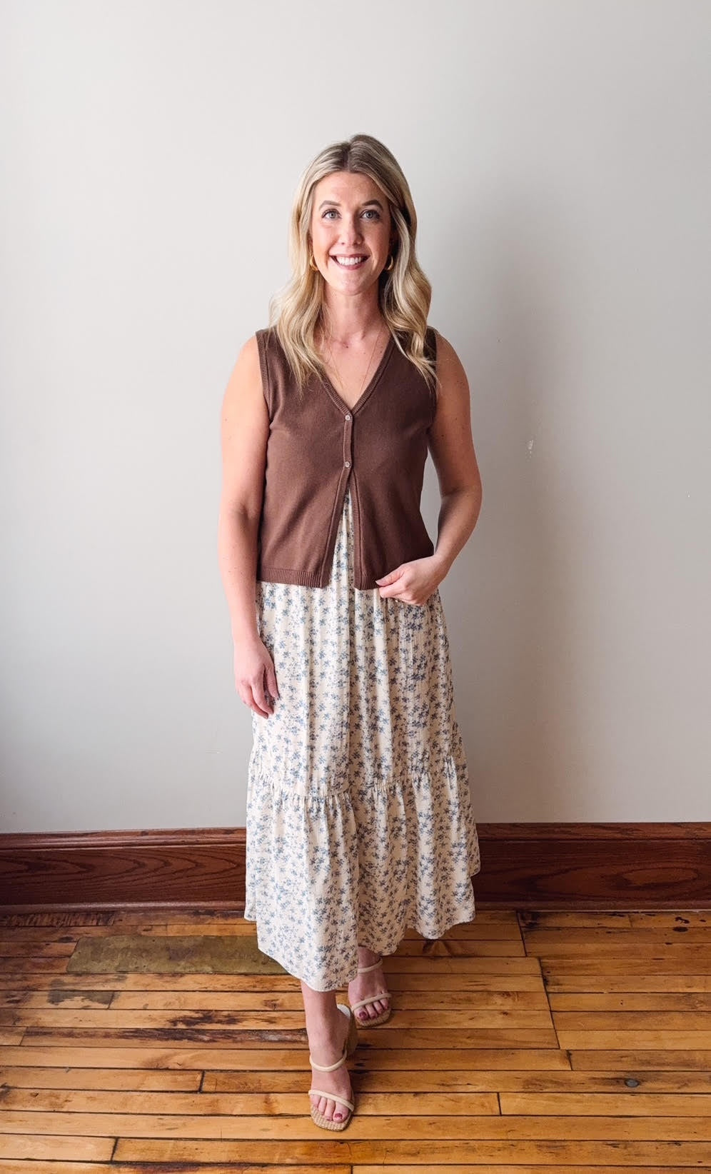 Woman wearing a brown sleeveless top and floral skirt standing on a wooden floor against a white wall.