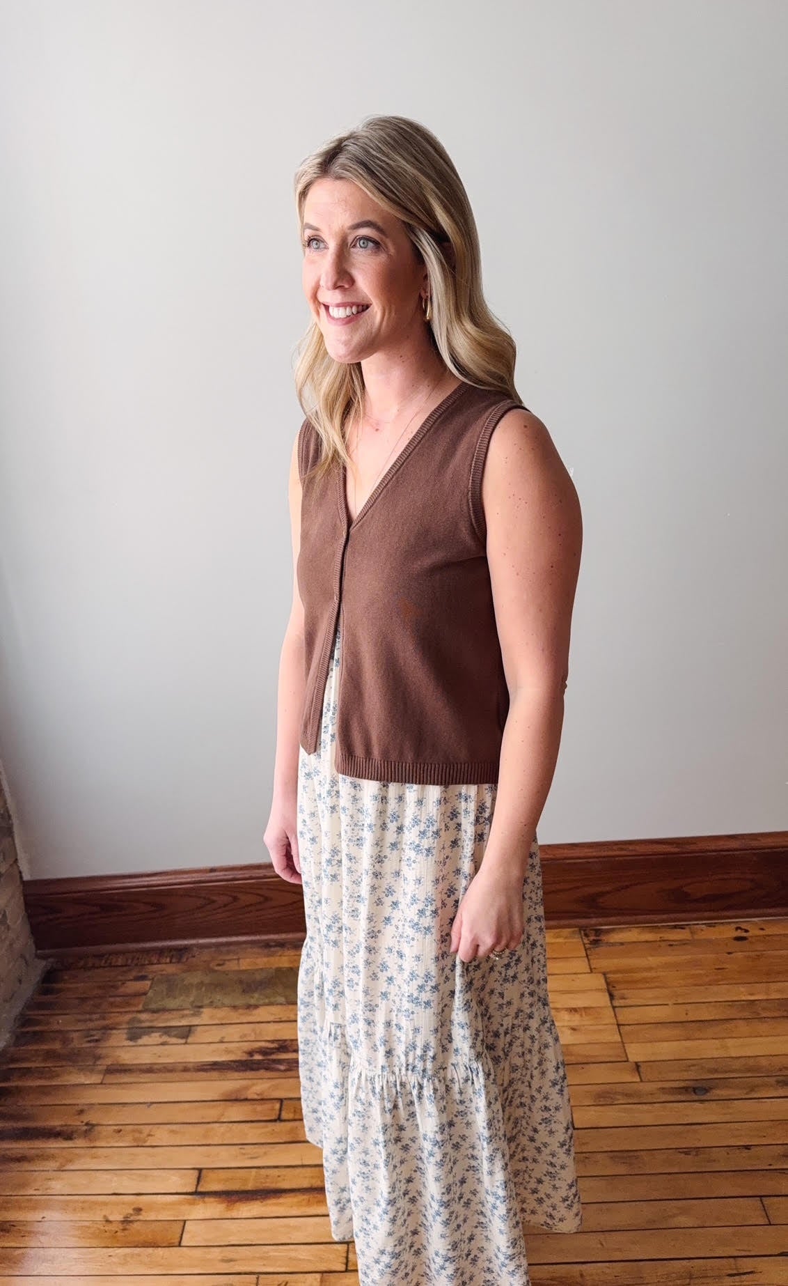 Woman wearing a brown sleeveless top and floral dress standing on a wooden floor.