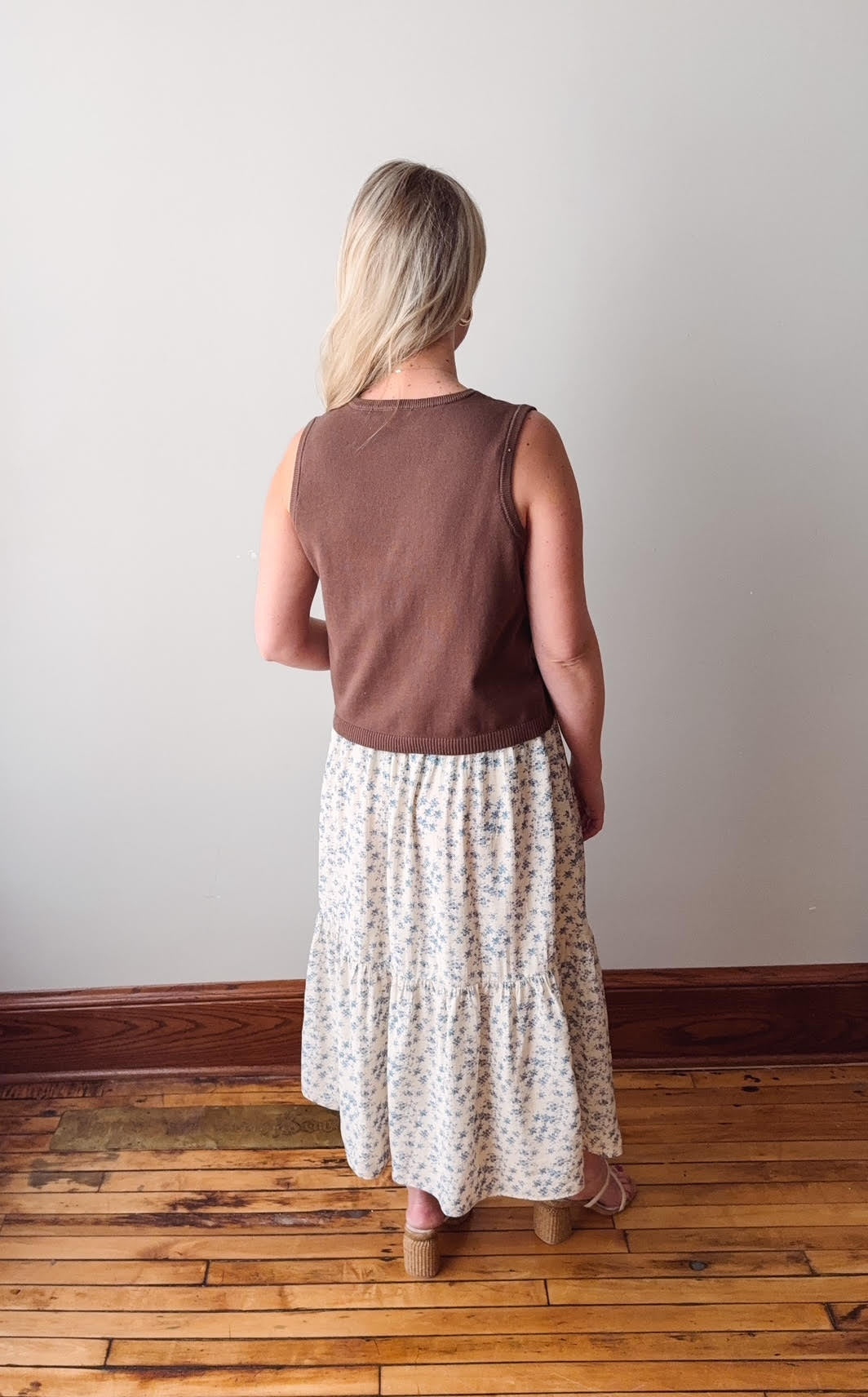 Person wearing a brown sleeveless top and white floral skirt standing on a wooden floor.