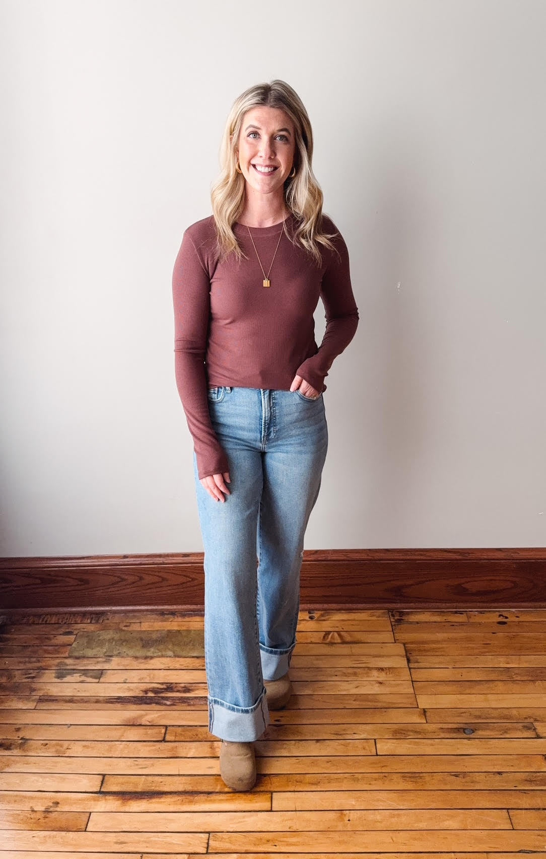 Woman wearing a maroon long-sleeve top and light blue jeans standing on a wooden floor.