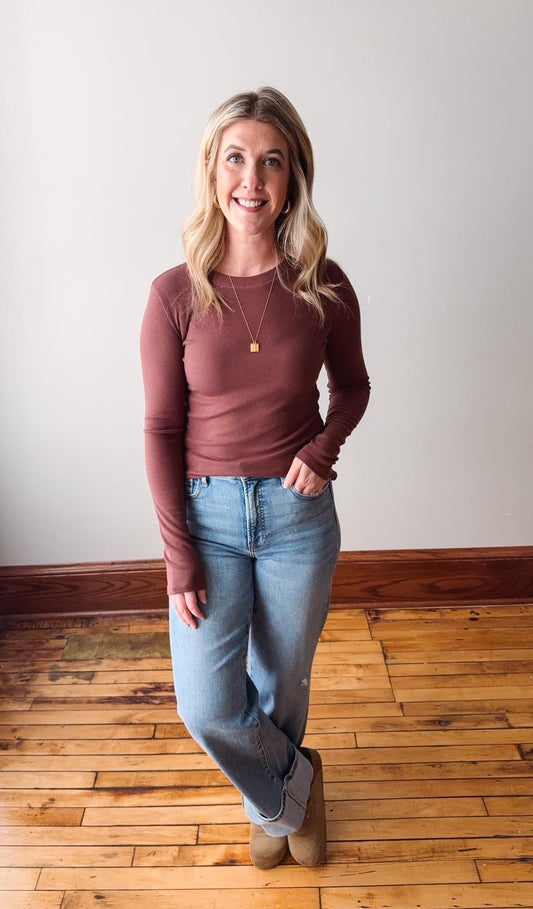 Woman wearing a brown long-sleeve top and blue jeans standing on a wooden floor.