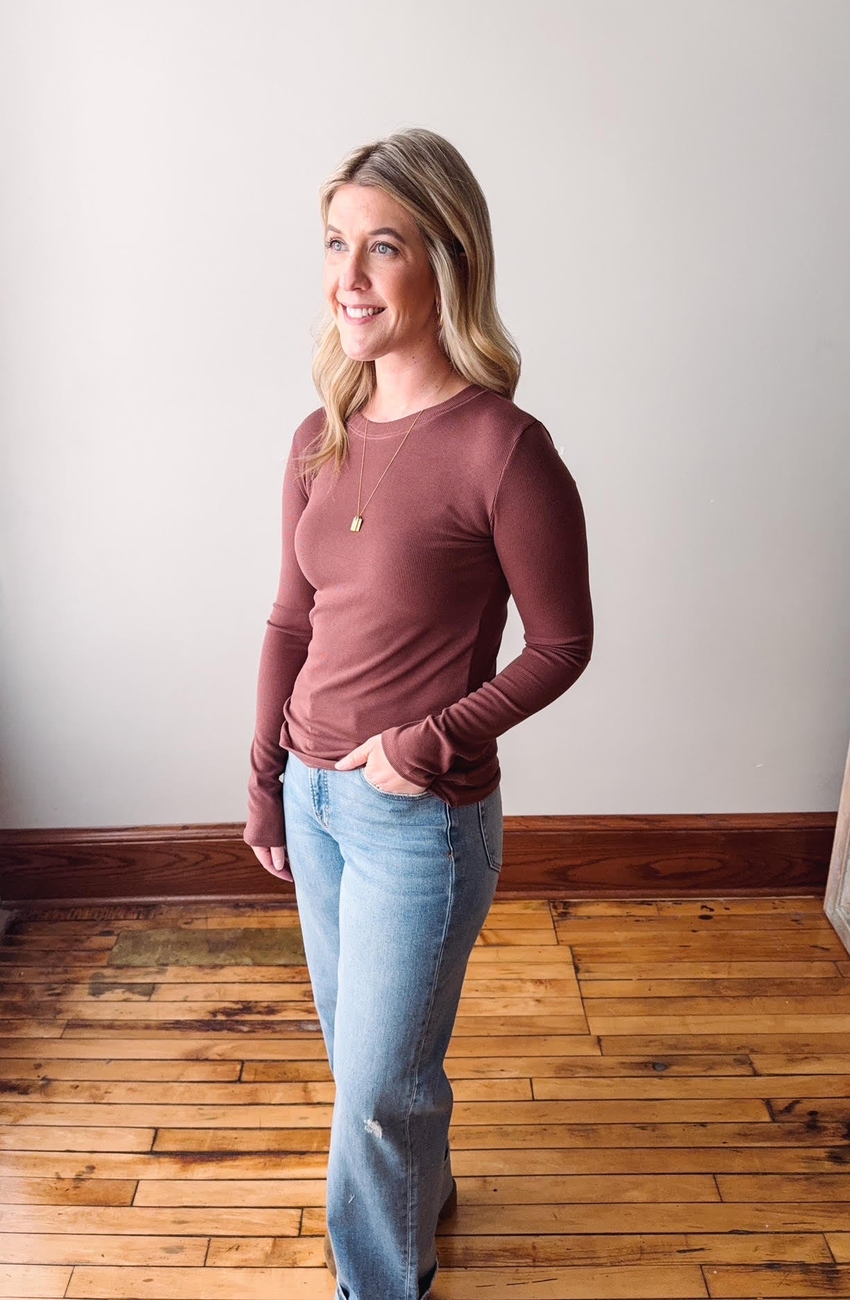Woman wearing a long-sleeve top and jeans standing on a wooden floor with a white wall background