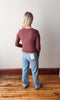 Person wearing a maroon long-sleeve top and blue jeans standing on a wooden floor with a white wall background.
