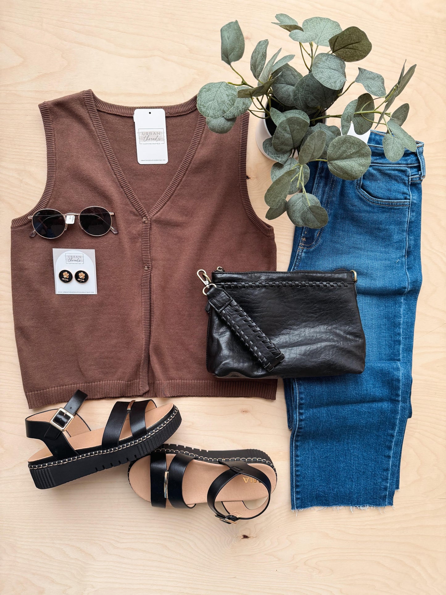 Brown sleeveless top, blue jeans, black sandals, black clutch, and sunglasses on a wooden surface with a plant.