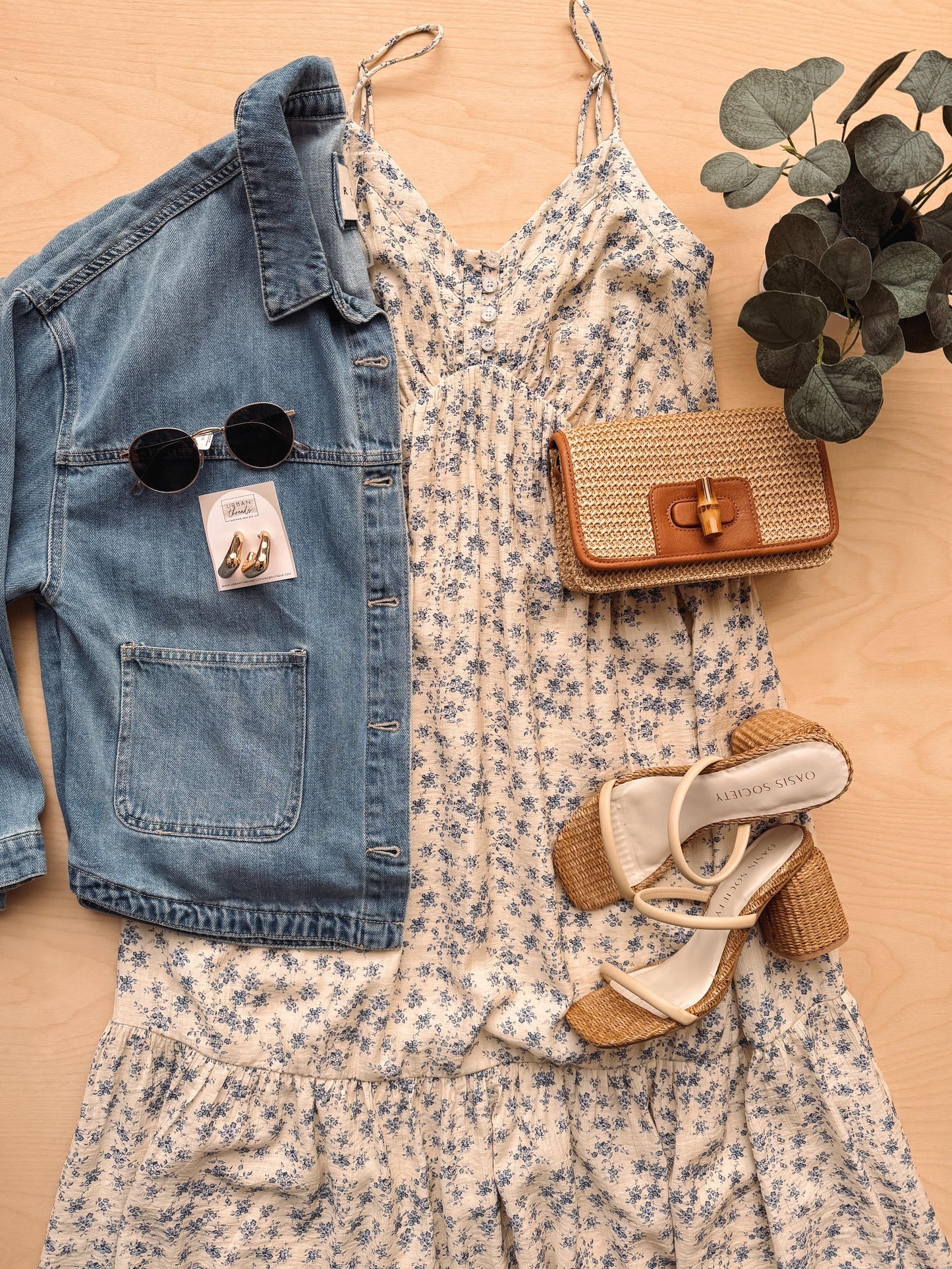 Denim jacket, floral dress, sunglasses, handbag, and shoes arranged on a wooden surface.