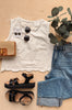 White sleeveless top, black sandals, blue jeans, and a woven handbag on a wooden surface with greenery.