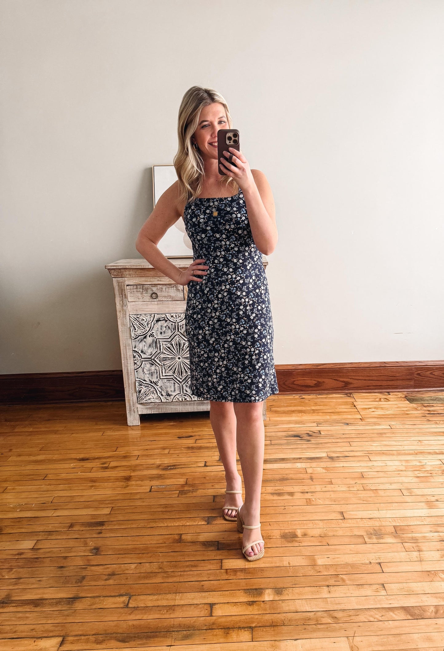 Woman taking a mirror selfie wearing a floral dress in a room with wooden flooring and a decorative cabinet.