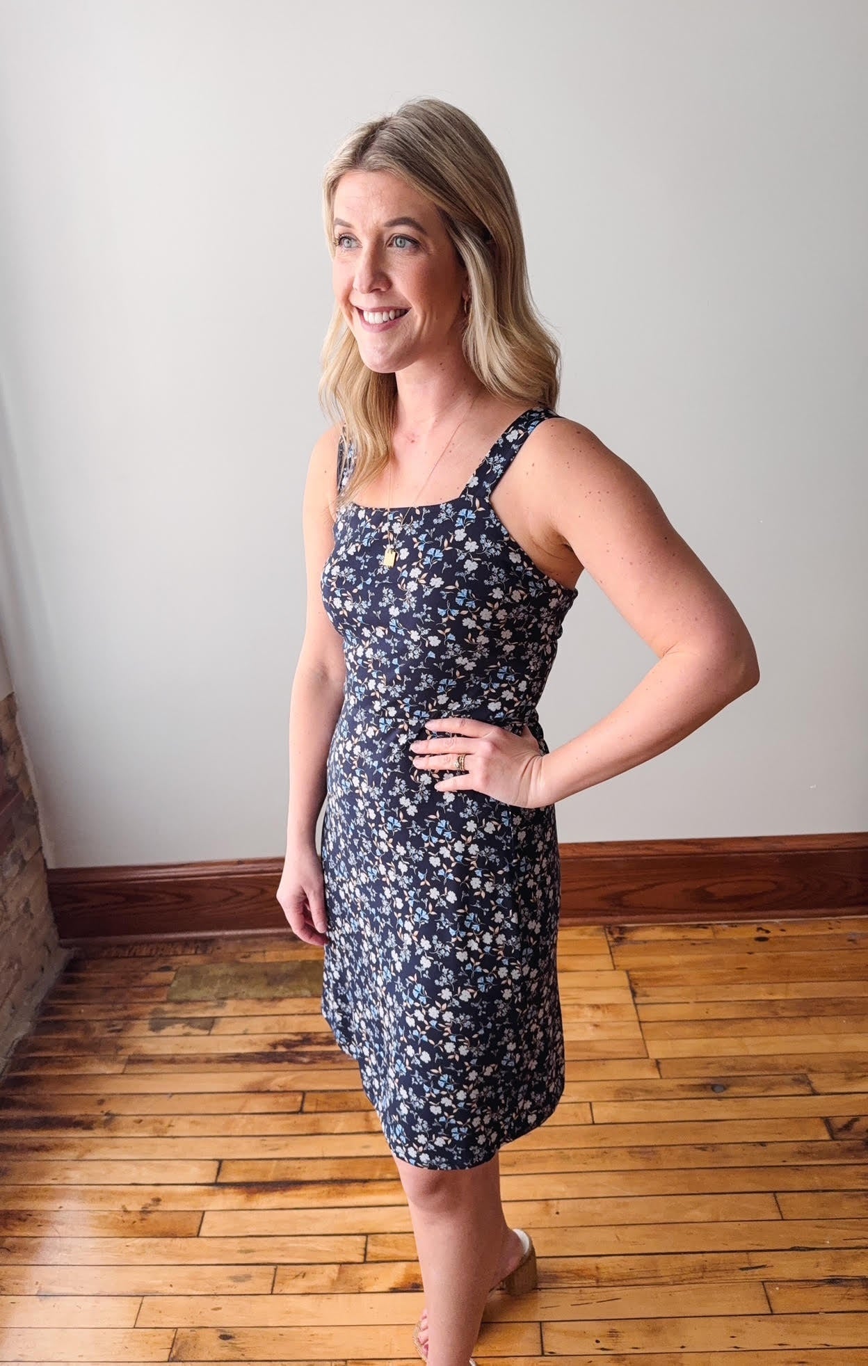 Woman wearing a navy floral dress standing in a room with wooden flooring and a white wall.