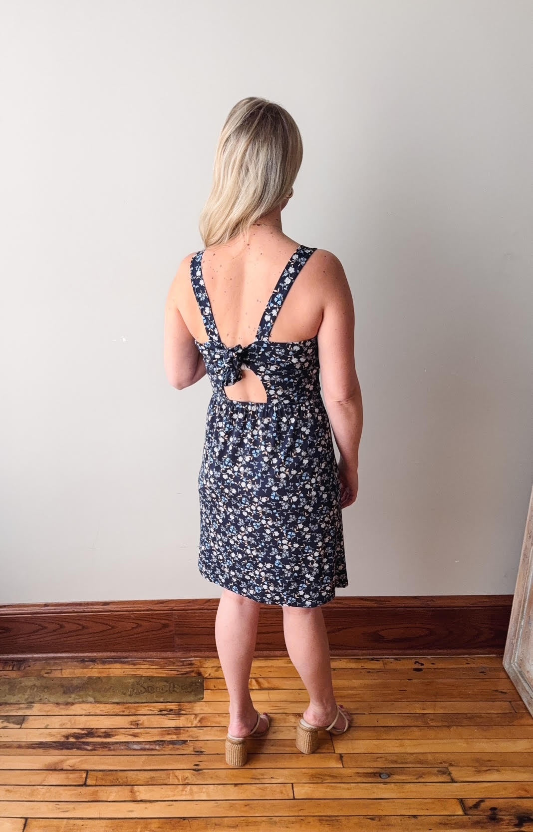 Woman wearing a floral dress with a tie back detail, standing on a wooden floor against a plain wall.