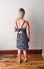 Woman wearing a floral dress with a tie back detail, standing on a wooden floor against a plain wall.