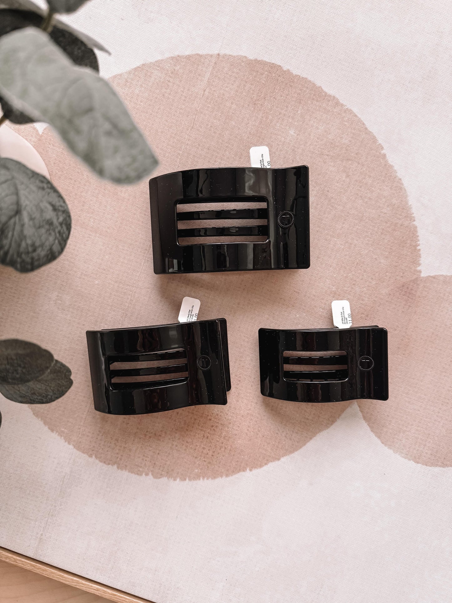 Three black hair clips on a pink and beige abstract background with green leaves.