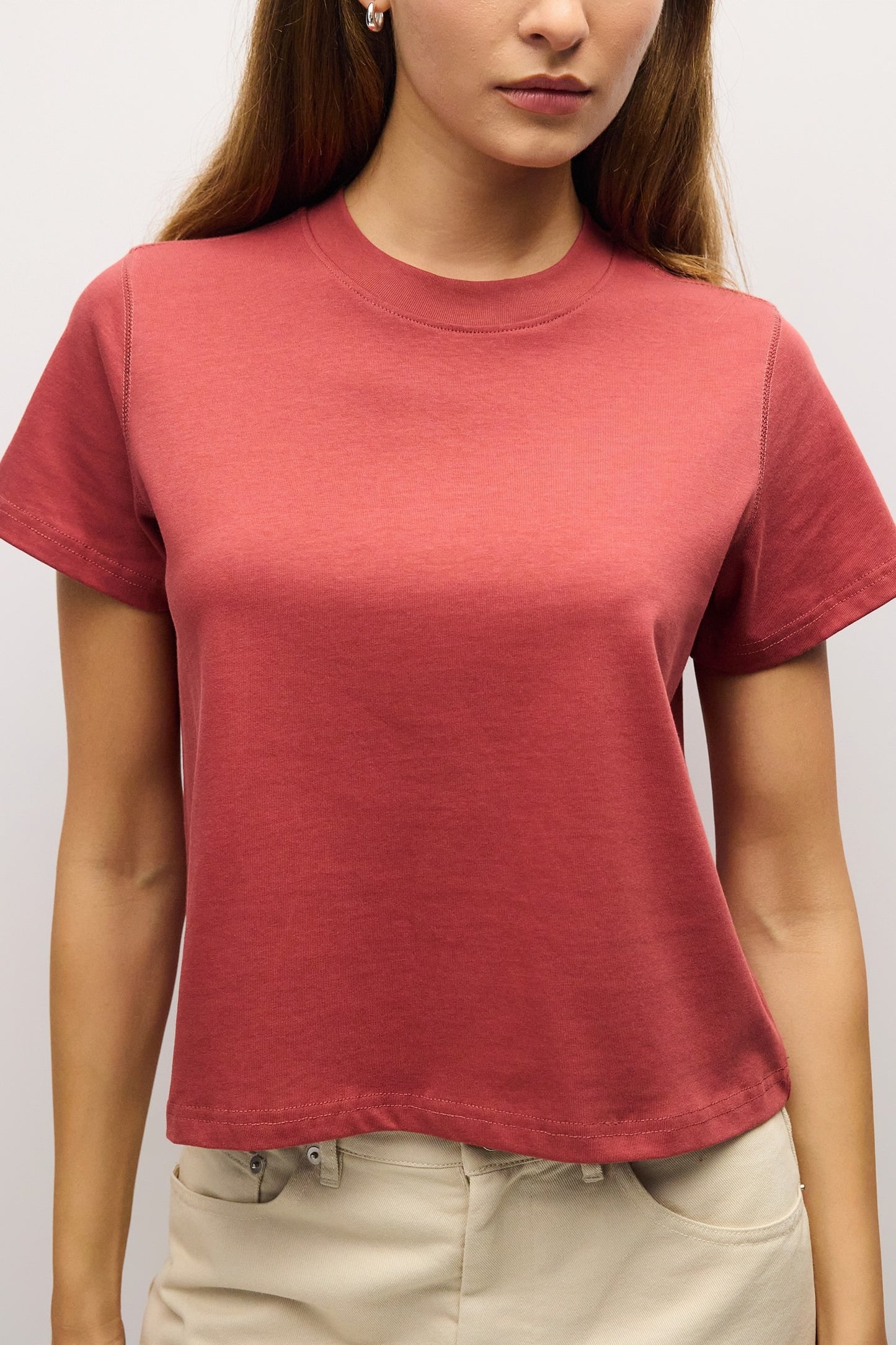 A woman wearing a short-sleeve crewneck t-shirt in a dusty rose or muted terracotta color, paired with beige pants, shown from the chest down against a plain light background.