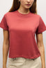 A woman wearing a short-sleeve crewneck t-shirt in a dusty rose or muted terracotta color, paired with beige pants, shown from the chest down against a plain light background.