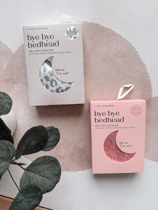 Two boxes of 'bye bye bedhead' baby satin pillowcases on a textured surface with green leaves.