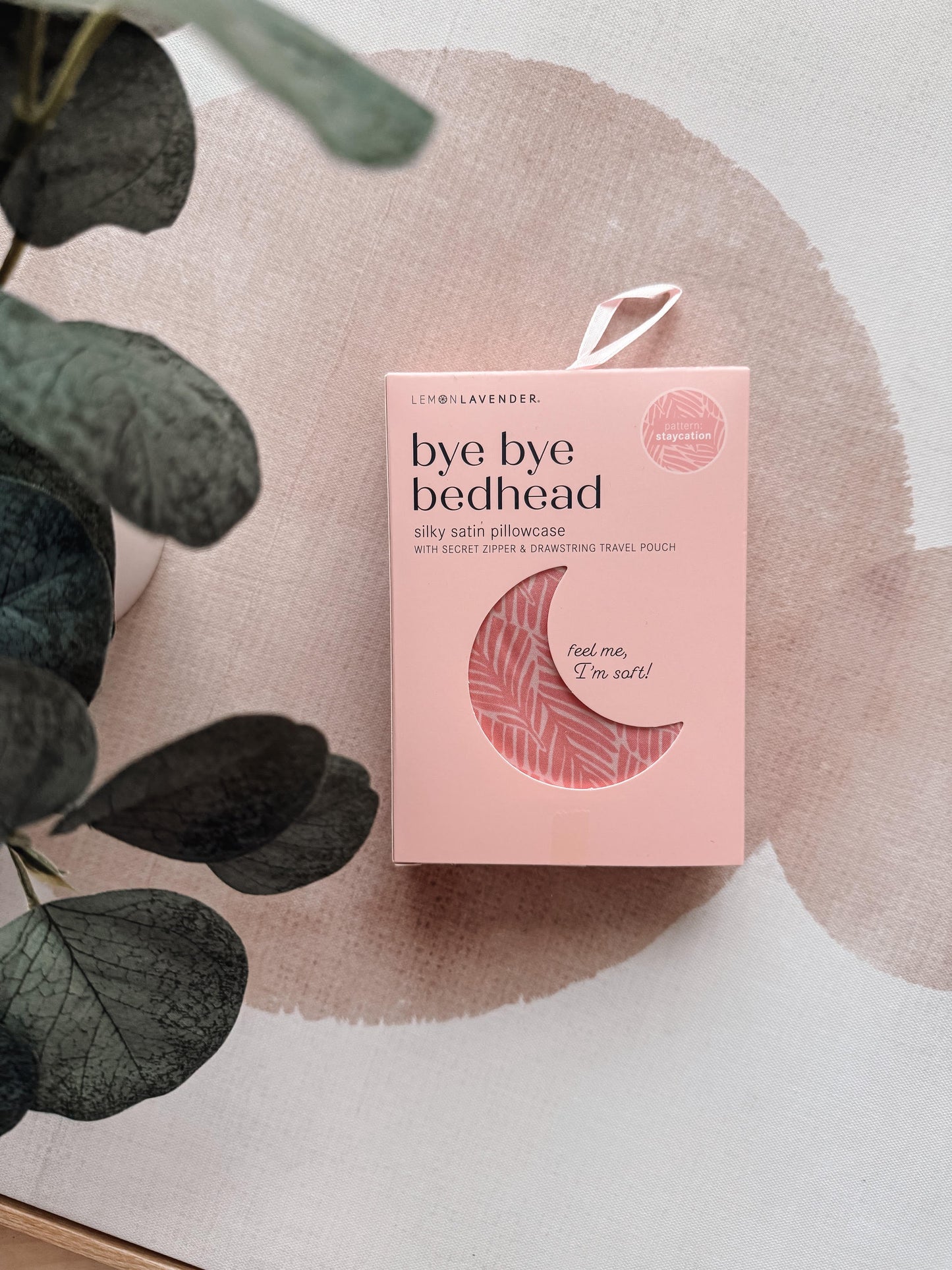 Pink 'bye bye bedhead' product packaging on a textured surface with green leaves.