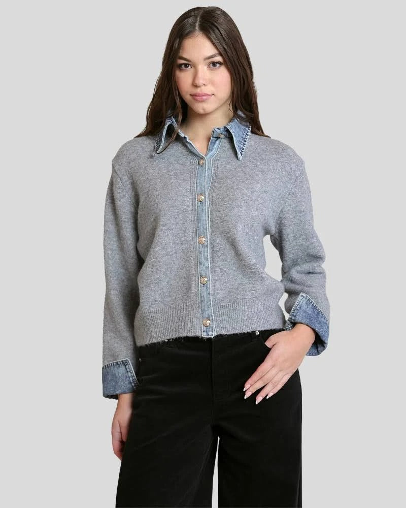 A woman modeling a cropped, light gray knit button-up shirt with a light blue denim collar, cuffs, and back placket, paired with black pants, against a solid light gray background.