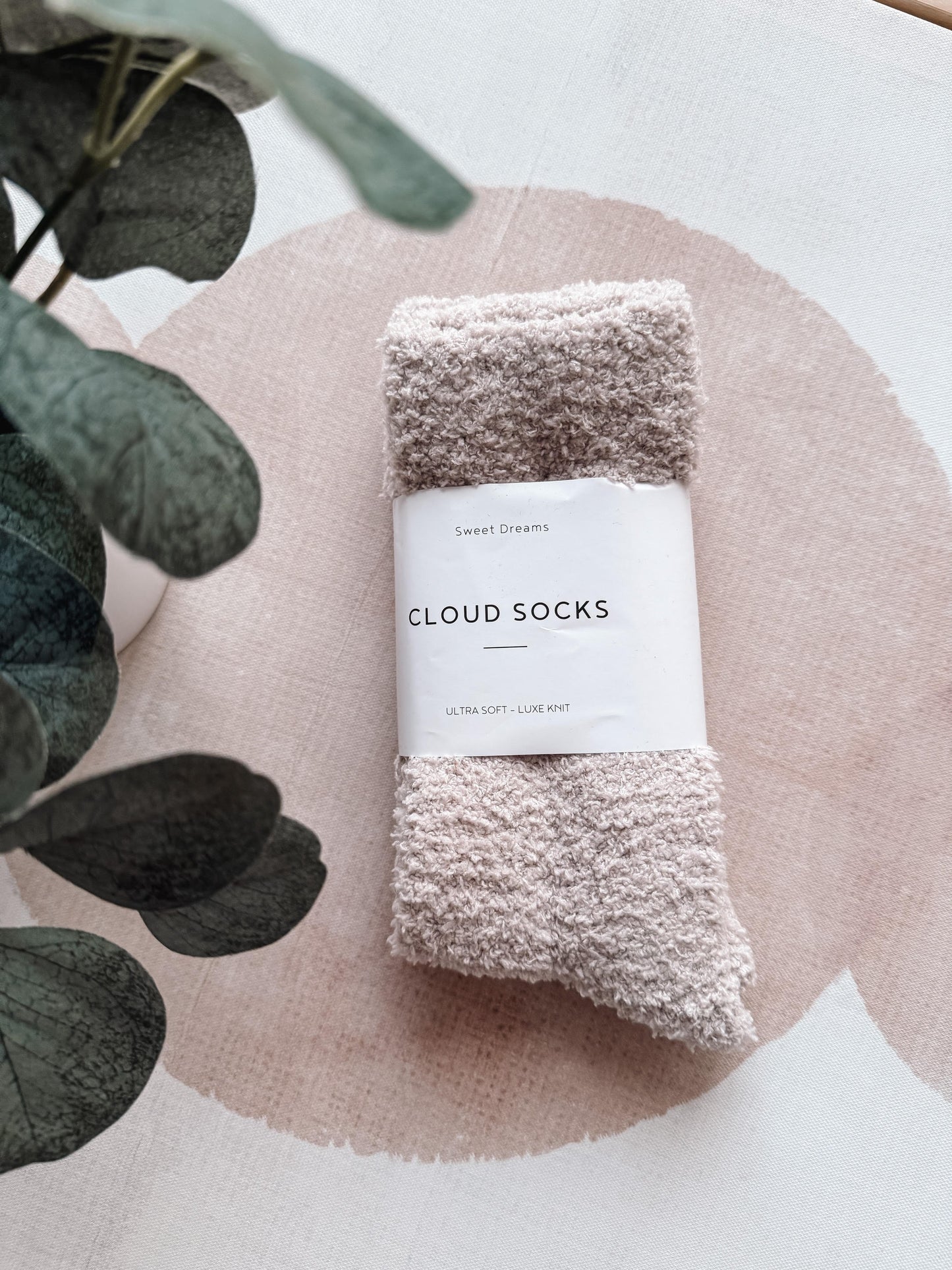 Pair of cloud socks with a label on a pink circular mat with eucalyptus leaves.