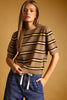 A woman wearing a brown short-sleeve knit sweater with horizontal beige and dark brown stripes, paired with blue jeans, posed against a solid warm brown background.