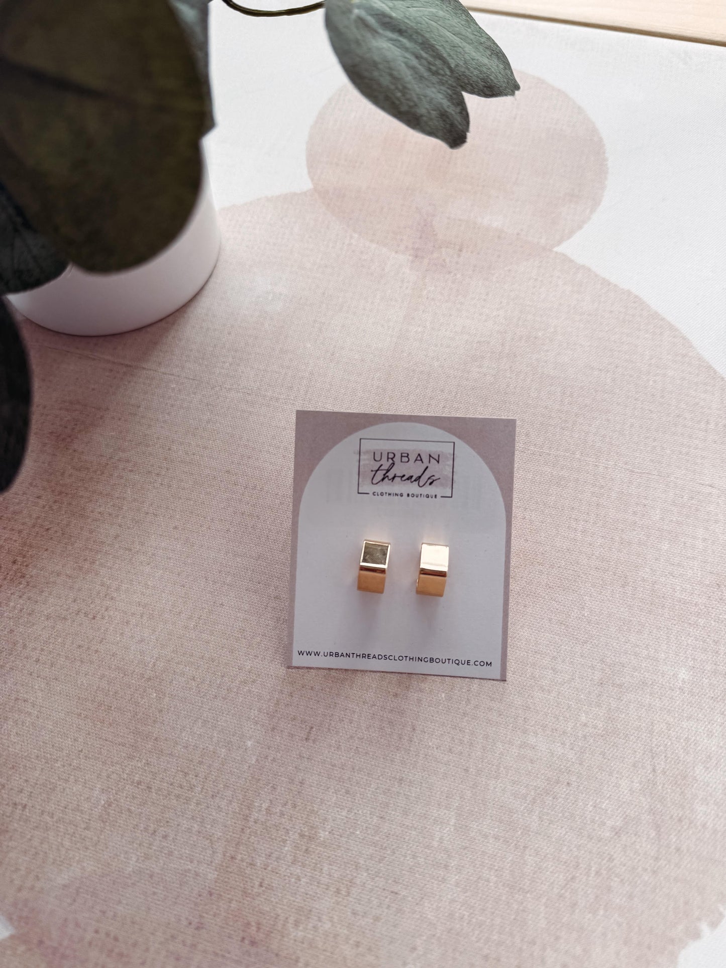 Gold square earrings on a branded card with a pink heart-shaped pillow in the background