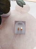 Gold square earrings on a branded card with a pink heart-shaped pillow in the background