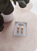 Set of gold earrings on a branded card with a blurred plant in the background