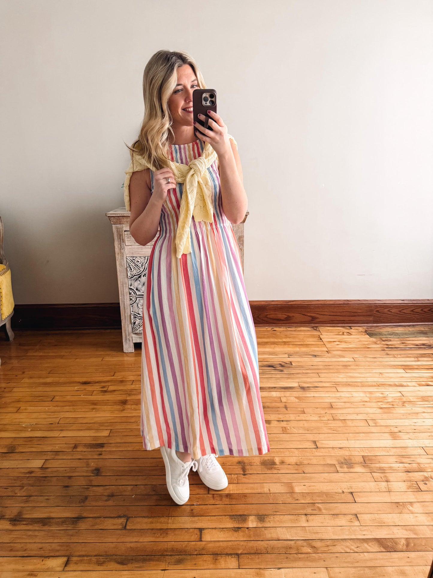 Woman taking a mirror selfie wearing a colorful striped dress and yellow scarf in a room with wooden flooring.