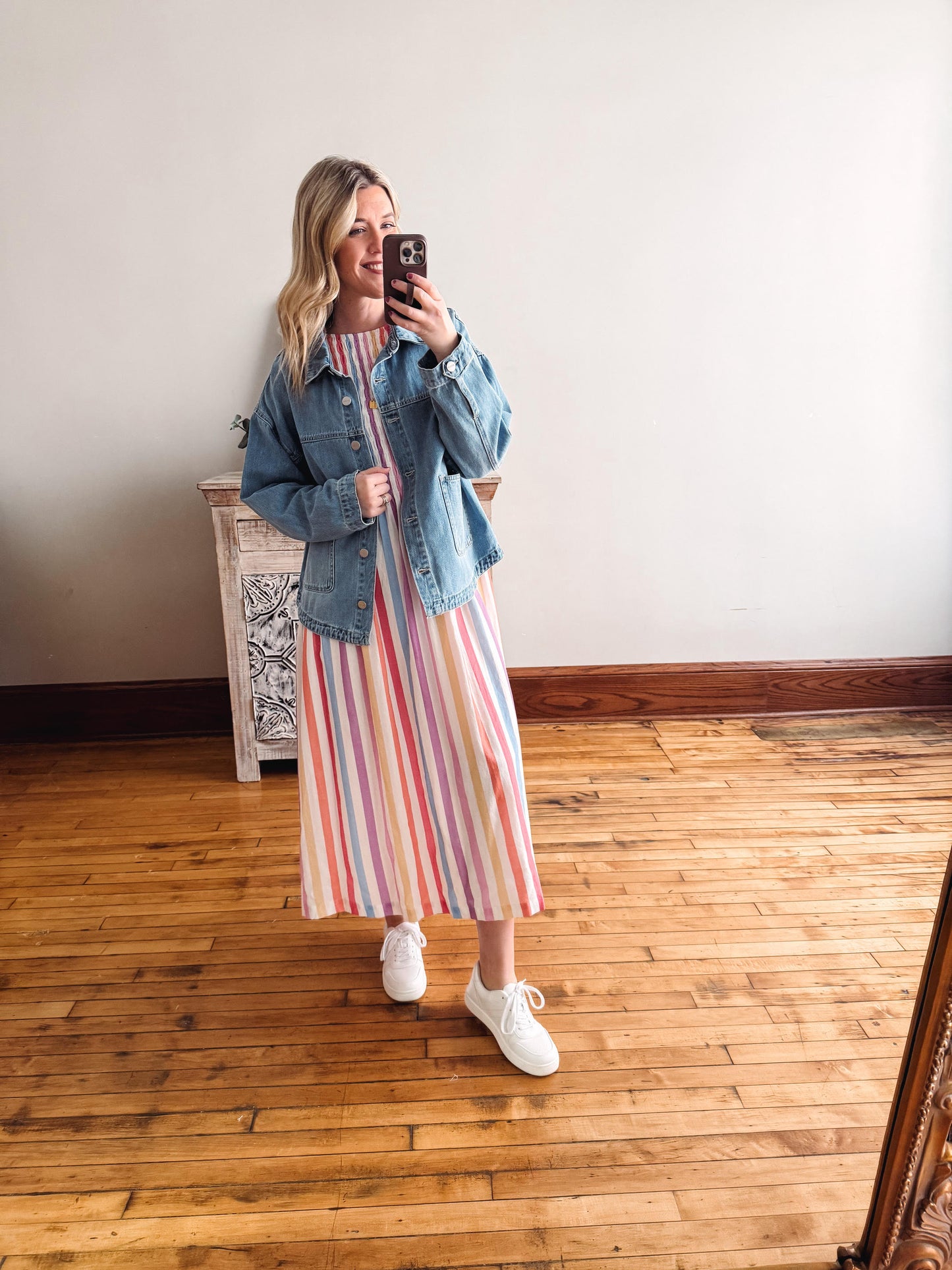 Woman taking a mirror selfie wearing a striped dress, denim jacket, and white sneakers on a wooden floor.