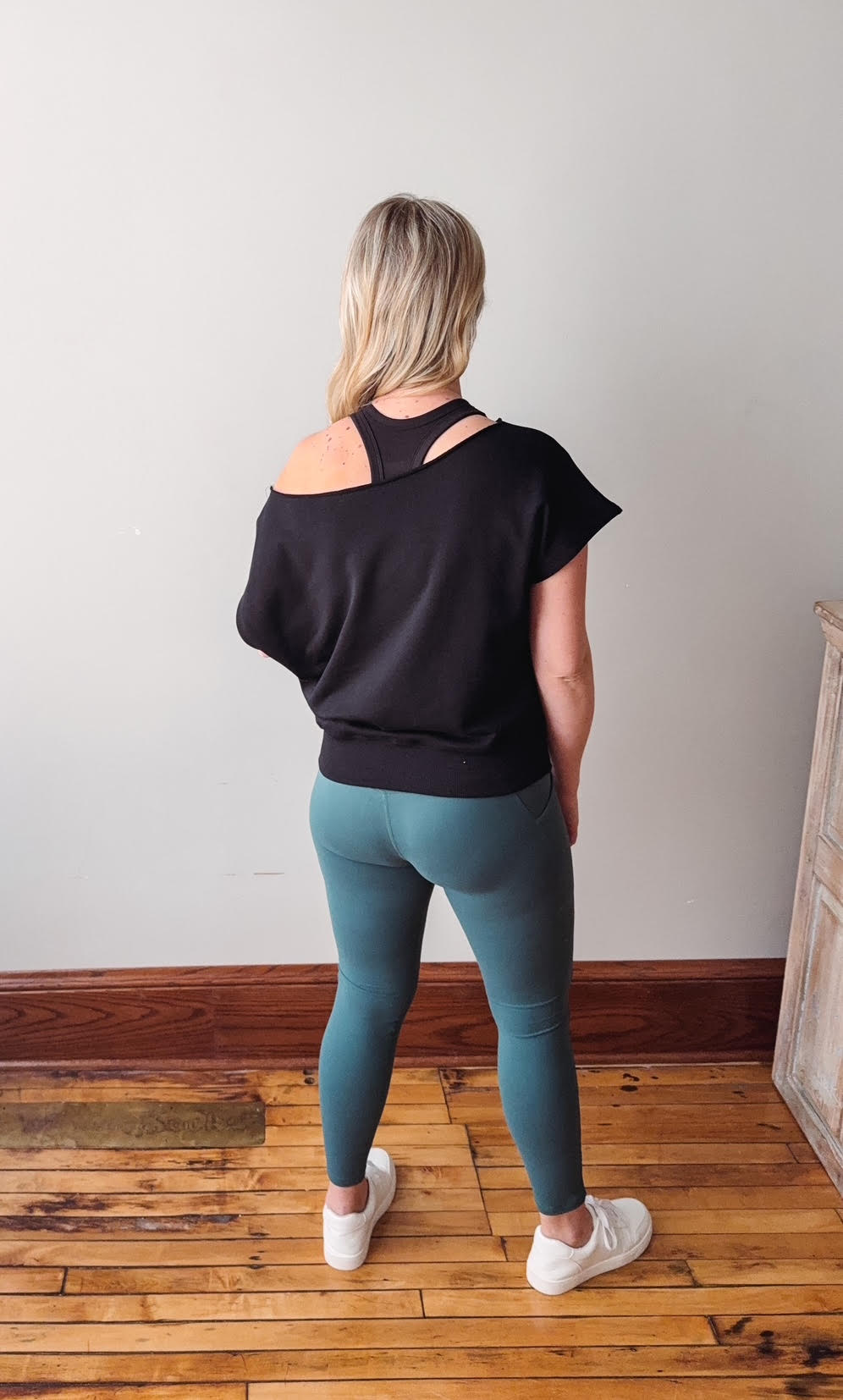 Woman wearing a black top and teal leggings standing on a wooden floor.