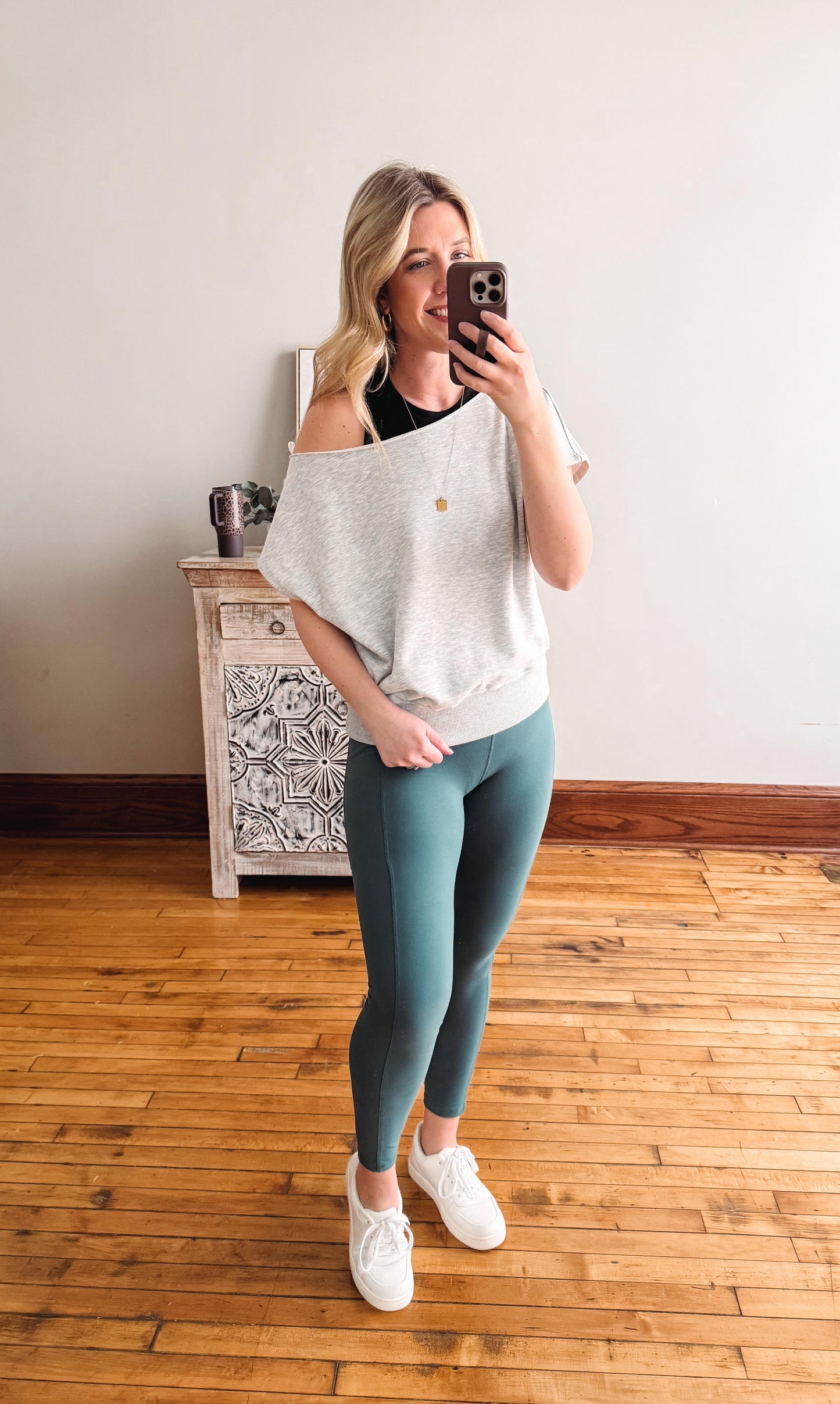 Woman taking a mirror selfie wearing a light gray off-shoulder top and teal leggings in a room with wooden flooring.
