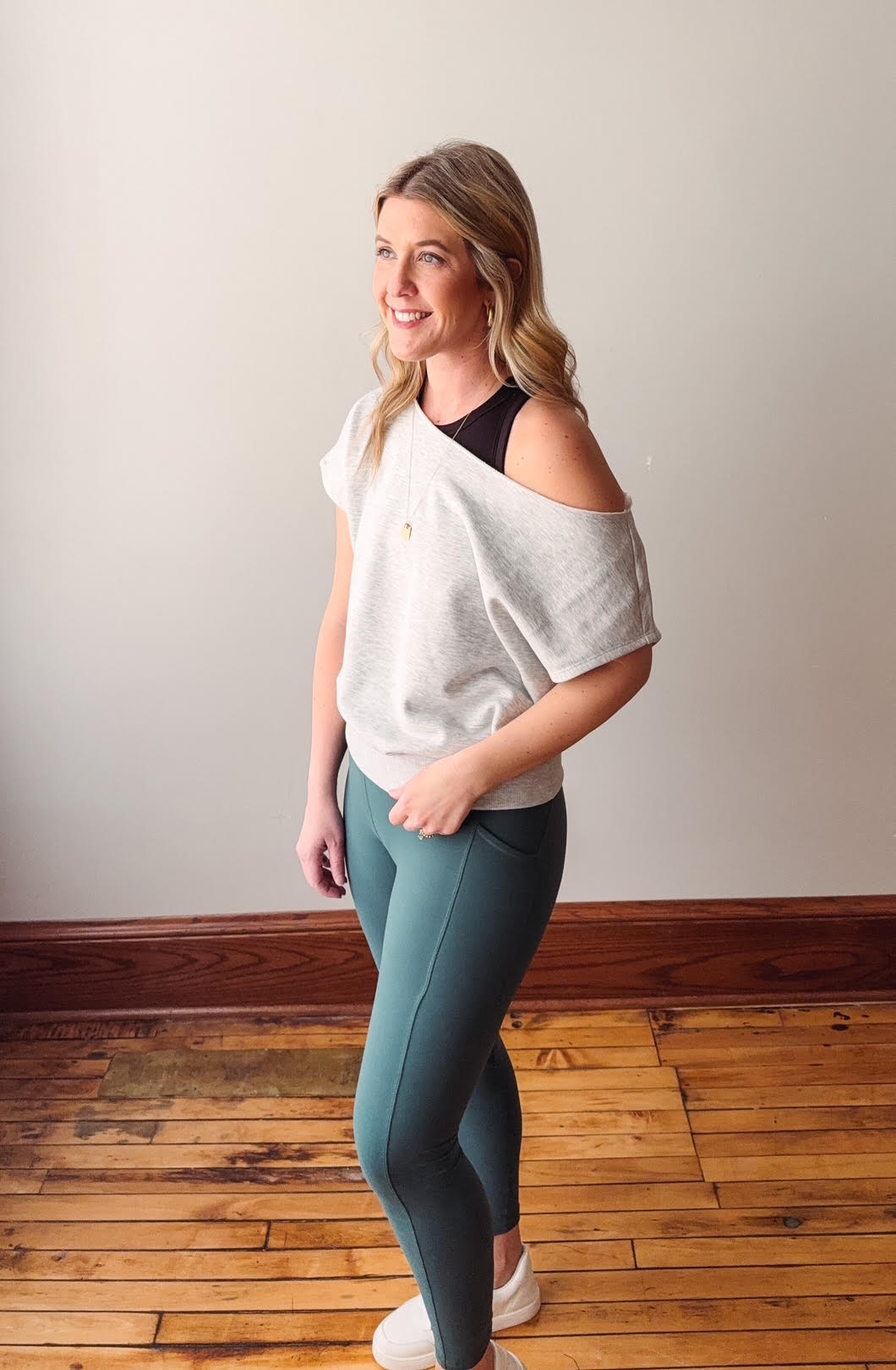 Woman wearing a white top and teal leggings standing on a wooden floor.