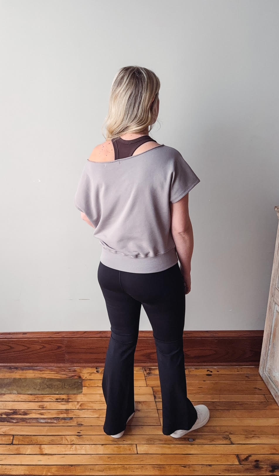 Person wearing a light gray top and black pants standing on a wooden floor with a white wall background