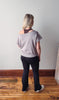 Person wearing a light gray top and black pants standing on a wooden floor with a white wall background