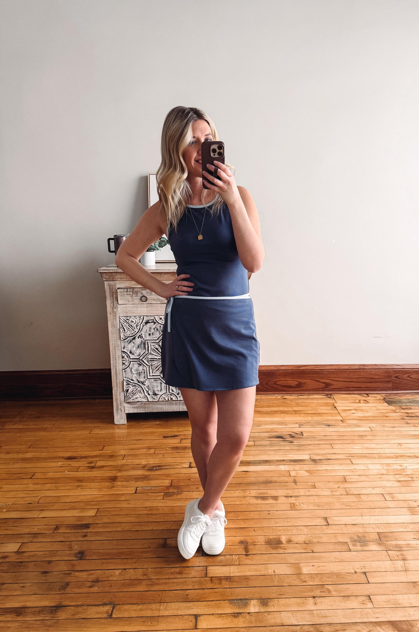 Woman taking a mirror selfie wearing a blue dress and white sneakers in a room with wooden flooring.