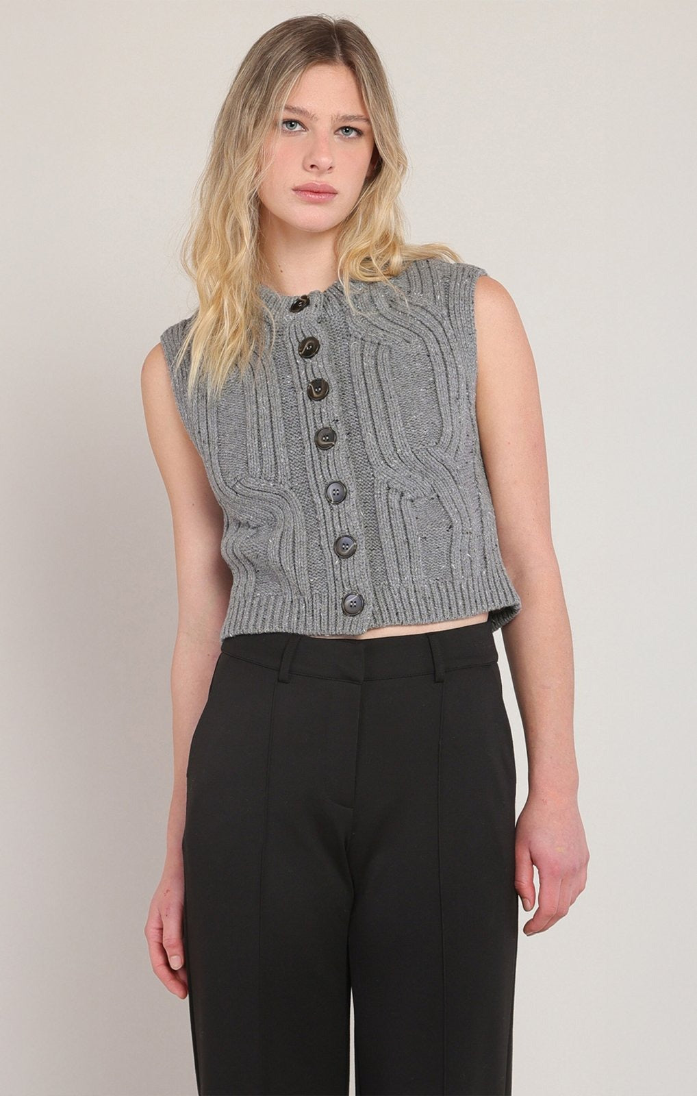 A woman modeling a sleeveless gray cable-knit cropped vest with dark buttons, paired with black trousers, against a plain light gray studio background.