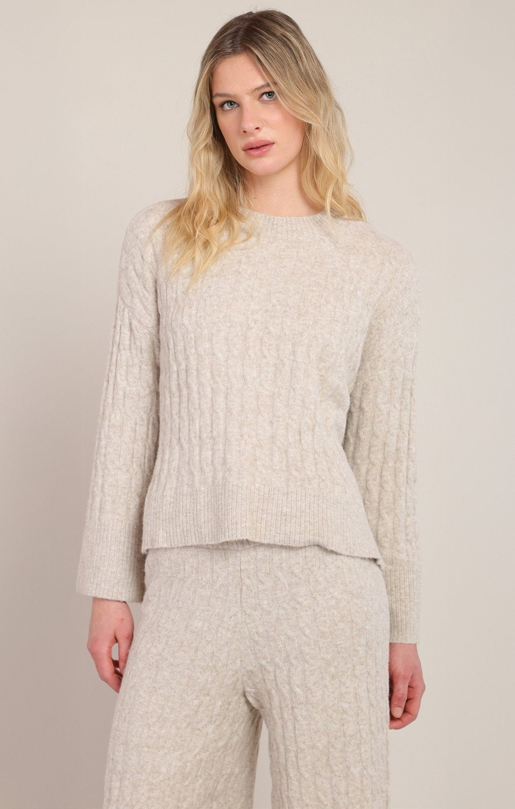 A woman modeling a light beige cable-knit sweater and matching knit pants against a plain light beige studio background.