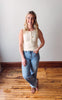 Woman wearing a beige sleeveless top and blue jeans standing on a wooden floor.