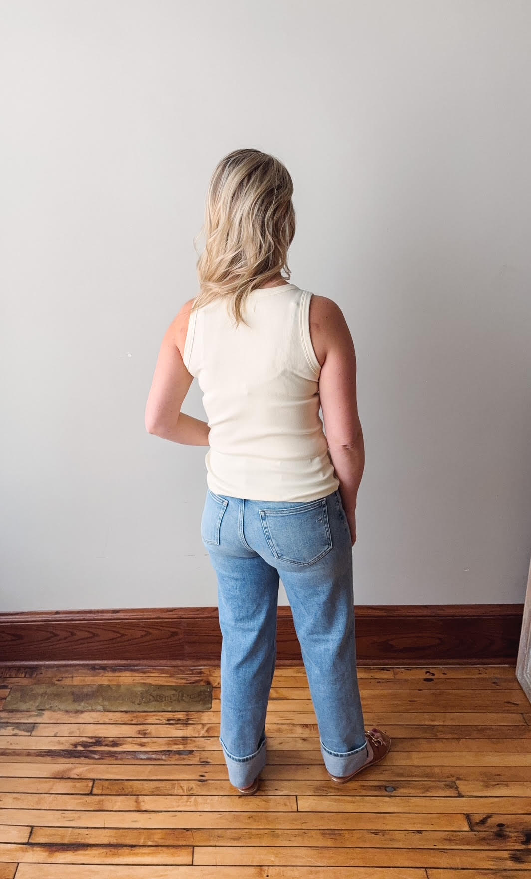 Person wearing a white tank top and blue jeans standing in an empty room with wooden flooring and a white wall.