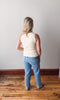 Person wearing a white tank top and blue jeans standing in an empty room with wooden flooring and a white wall.