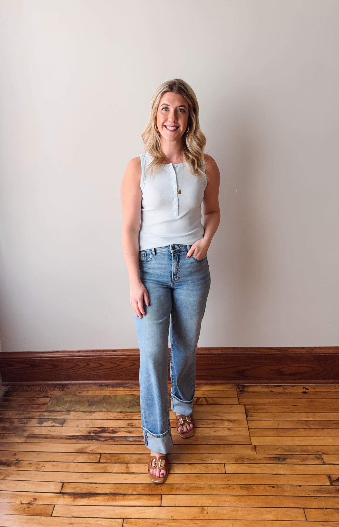 Woman wearing a light blue sleeveless top and jeans standing on a wooden floor with a white wall background.