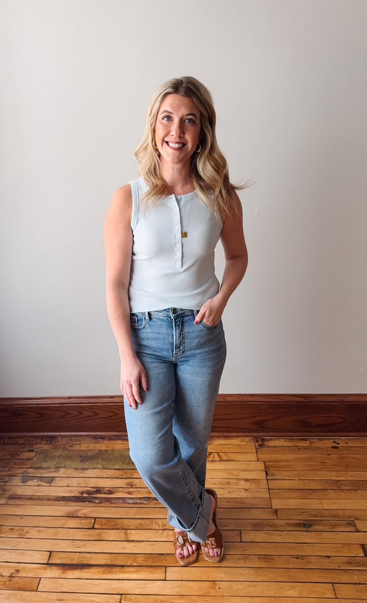 Woman wearing a light blue sleeveless top and jeans standing on a wooden floor.
