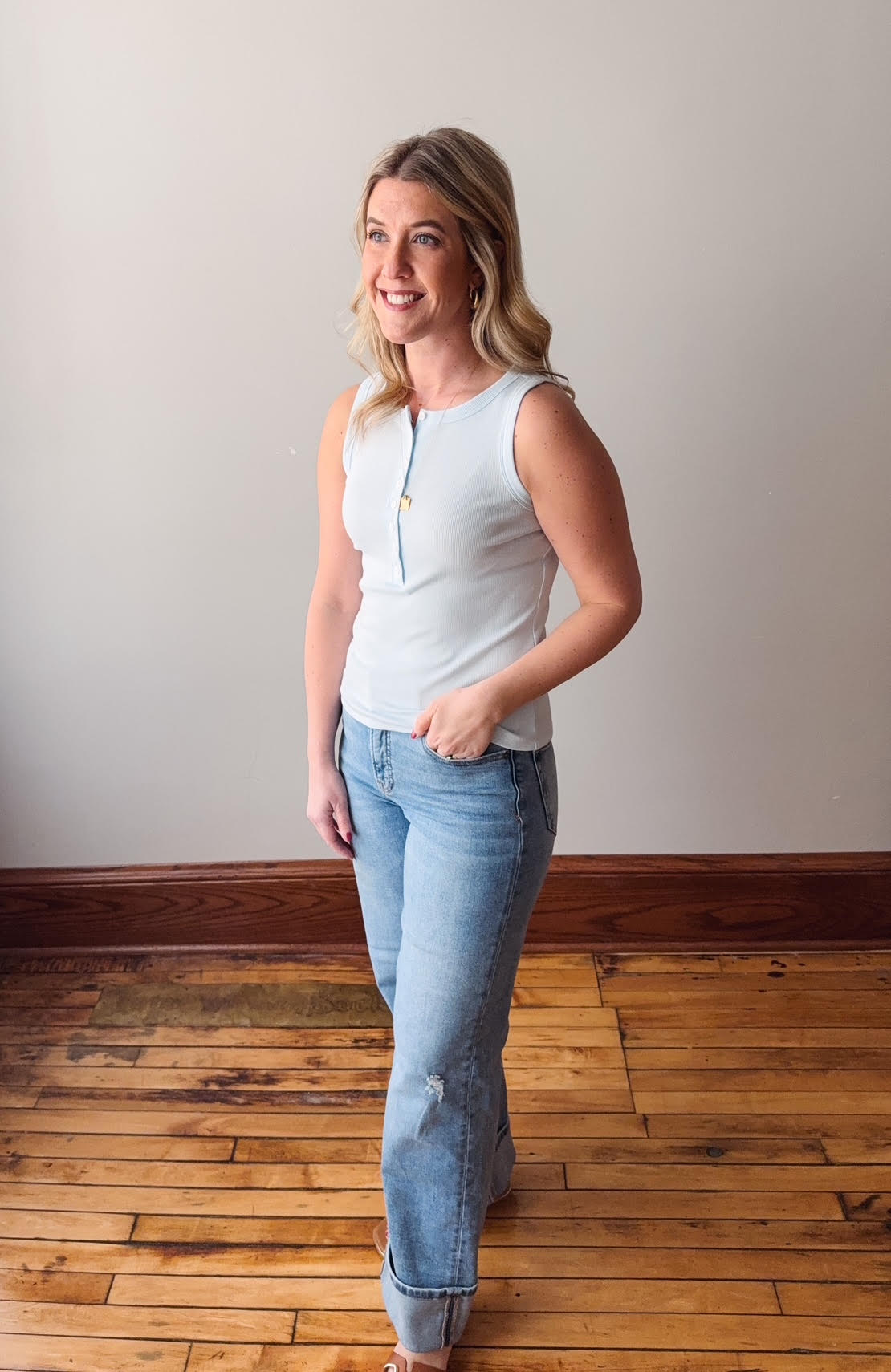 Woman wearing a light blue sleeveless top and jeans standing on a wooden floor.