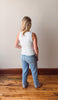 Person wearing a white tank top and blue jeans standing on a wooden floor with a plain wall background
