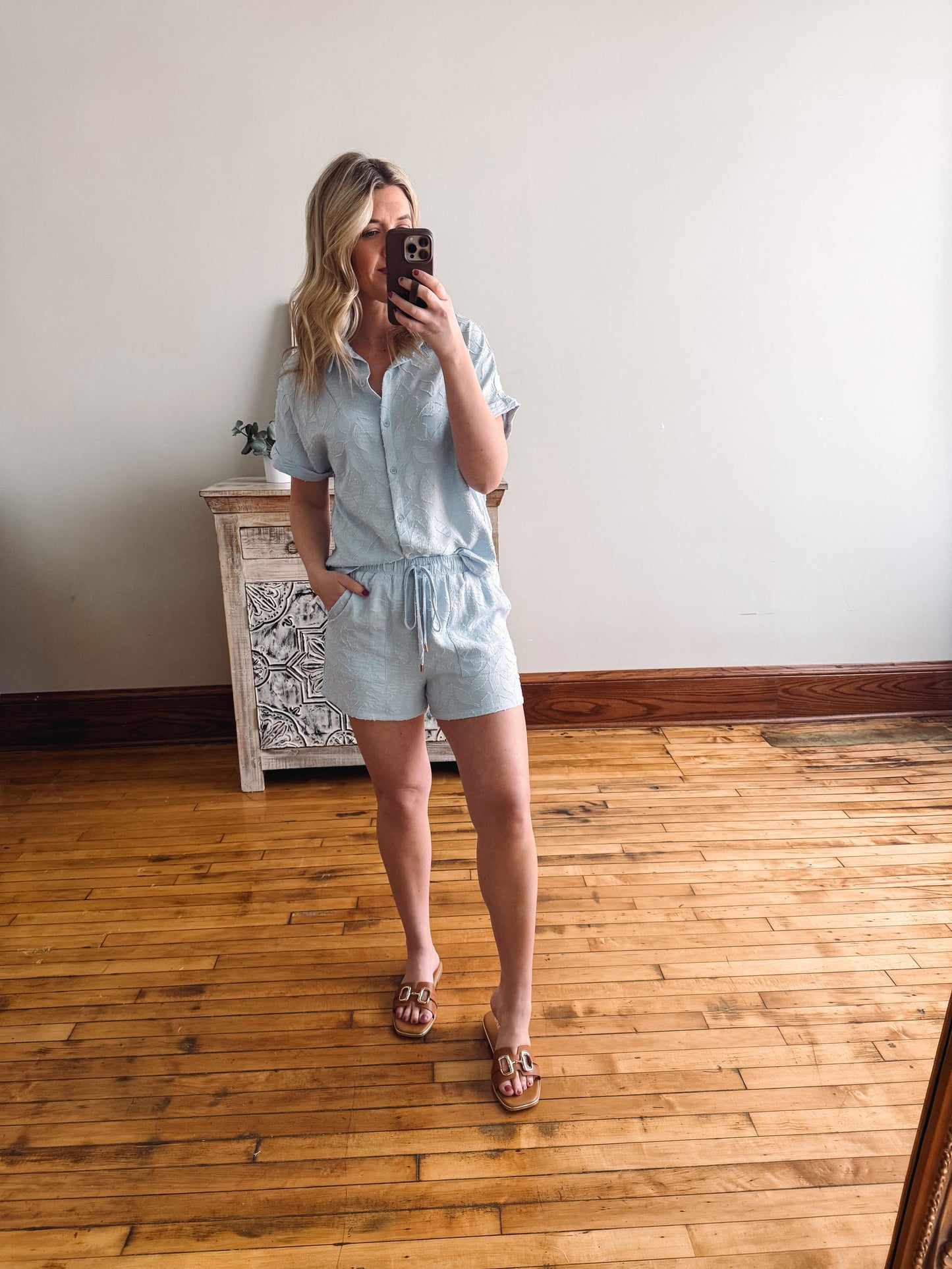 Woman taking a mirror selfie wearing a light blue button-up shirt and shorts in a room with wooden flooring.